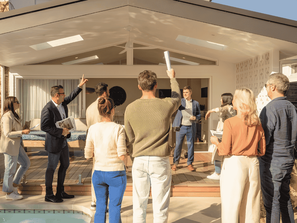 Group of people at an outdoor house tour listening to a presenter who raises a rolled document on a modern patio.