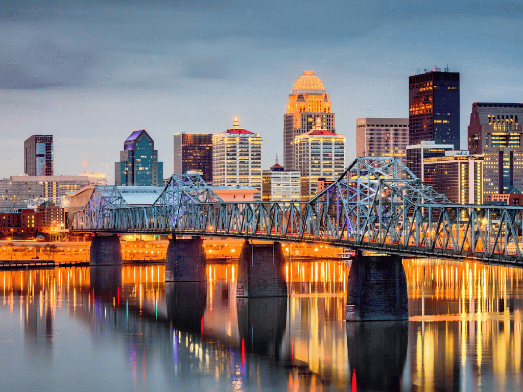 Evening city skyline with a blue steel truss bridge crossing a reflective river and warm lights. deep blue sky and illuminated buildings in the background.